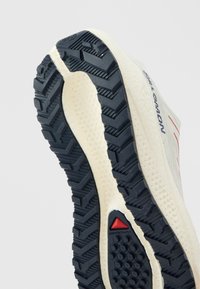 Valge ja must jooksujalatsi tald sügava mustriga, punane kolmnurkne logo ja "Salomon" bränd külje peal.