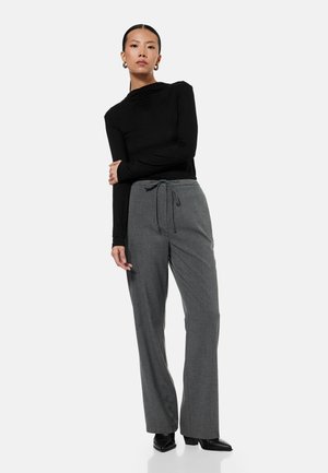 Nainen seisoo käsivarret ristissä, päällään musta pitkähihaisen paita, harmaat nyöripöksyt ja mustat teräväkärkiset kengät valkoisella taustalla.