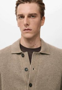 Beige gebreid vest met een kraag, voorzien van vier zwarte knopen en een subtiel textuurpatroon. Gedragen over een donker shirt.