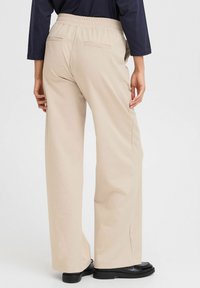 Beige vida byxor med elastisk midja, slät yta och två bakfickor; tillsammans med svarta skor för en komplett look.