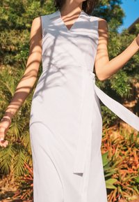 Vit, ärmlös omslagsklänning med djup V-ringning, detalj med sidoknytning, slät textur och flödande silhuett, mot en grön bakgrund.