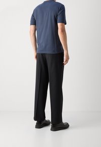 Mies, der ein navyblaues Kurzarmhemd, schwarze weite Hosen und schwarze Dressschuhe trägt, steht vor einem schlichten weißen Hintergrund.