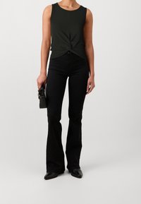 Top sin mangas de color verde oscuro con un diseño frontal retorcido, combinado con unos vaqueros negros de tiro alto y campana, y zapatos negros, sosteniendo un pequeño bolso negro.