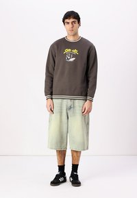 Felpa marrone con strisce bianche, dotata di stampa grafica e logo. Pantaloni corti in denim azzurro che arrivano fino al ginocchio. Sneakers nere con tre strisce.