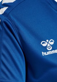 Maillot de sport bleu avec des bandes blanches en chevrons sur l'épaule et un logo d'abeille blanc avec le texte "hummel" sur la poitrine.