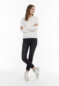 Fehér kötött pulóver, fekete slim fit farmernadrág és több színű sportcipő, texturált talppal. A modell lazán áll, karjai keresztbe tett helyzetben.