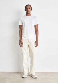 Weißes T-Shirt mit kurzen Ärmeln und Tasche, kombiniert mit cremefarbenen weit geschnittenen Hosen und grauen Sneakers, vor einem neutralen Hintergrund stehend.