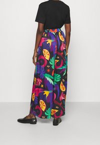 Personne portant un haut noir et un pantalon large coloré avec un motif de fruits tropicaux et de feuilles, se tenant devant un fond uni.