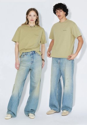 Deux personnes portant des t-shirts beige et des jeans bleus délavés à jambes larges se tiennent devant un fond blanc.