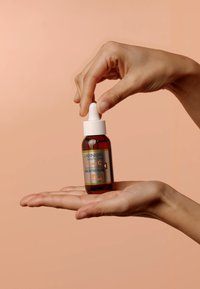 Bouteille en verre brun avec compte-gouttes blanc, étiquetée "Sérum de Nuit Éclaircissant à la Vitamine C Garnier SkinActive." Main tenant la bouteille sur un fond pêche.