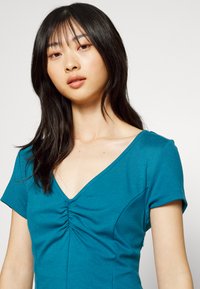 Top ajustado em verde azulado com mangas curtas, decote em V profundo e detalhe franzido no peito. Material de algodão suave, costuras mínimas e contornos subtis.