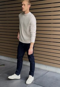 Lyshåret mand iført en lysegrå sweater, mørke jeans og hvide sneakers stående mod en brun væg med lameller.
