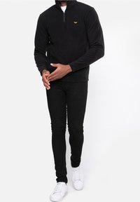 Pull-over en polaire noire à demi-fermeture éclair avec manches longues et poches latérales, associé à un jean noir ajusté et des baskets blanches. Détail notable du logo jaune.