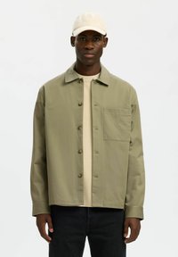 Homme portant une casquette beige, une veste kaki clair boutonnée avec une poche poitrine, un pull crème et un pantalon noir, debout devant un fond uni.