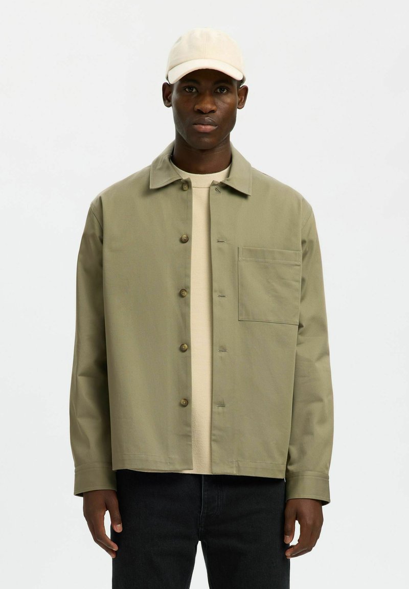 Homme portant une casquette beige, une veste kaki clair boutonnée avec une poche poitrine, un pull crème et un pantalon noir, debout devant un fond uni.