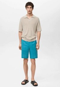 Polo shirt in maglina beige abbinato a pantaloni corti teal. I sandali hanno un design con una cintura nera. Texture lisce e una vestibilità rilassata in mostra.