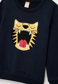 Sudadera azul marino con un gráfico de cara de gato amarillo que presenta una boca rugiente, rayas negras texturizadas y detalles de lengua roja.
