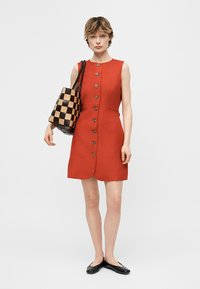 Femeie purtând rochie roșie fără mâneci, cu nasturi în față, ținând o geantă mare de tip tote, pătrată, maro și neagră, purtând pantofi negri comozi, stând pe fundal alb.