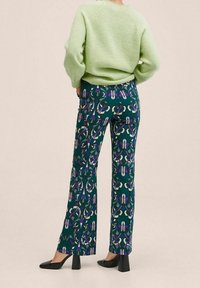 Zielony sweter o fakturze, szerokie spodnie w ciemnozielonym kolorze z abstrakcyjnym wzorem w odcieniach fioletu i kremu oraz czarne buty na słupku.