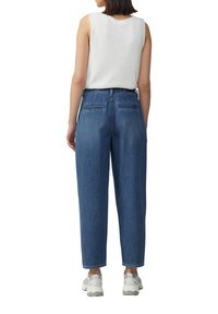 Jean bleu taille haute avec une coupe ample et des poches arrière, associé à un haut sans manches de couleur claire. Le mannequin porte des baskets blanches.