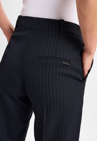 Close-up van een persoon die donkerblauwe gestreepte broek draagt met een achterzak en een hand in de zijzak, gecombineerd met een witte geribde top.