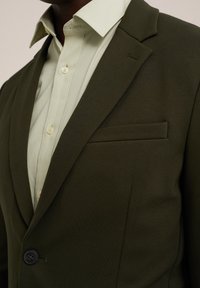 Blazer verde olivo con una textura suave, cierre de un botón y un bolsillo en el pecho, complementado con una camisa abotonada verde claro debajo.
