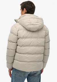 Kurtka puffer w jasnym beżu, z kapturem, pikowanym wzorem i elastycznymi mankietami. Z tyłu widoczna jest gładka tekstura z minimalnymi detalami.