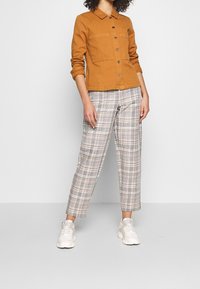 Světle hnědá košile na knoflíky s dvěma kapsami na hrudi, v kombinaci se šedými kostkovanými kalhotami s různobarevnými pruhy a bílými teniskami.