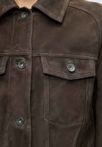Gros plan sur une veste en daim brun foncé avec poches avant et boutons en métal gravés du logo "DRYKORN".