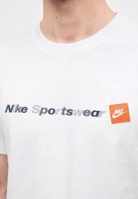 Κοντινή λήψη ενός λευκού T-shirt Nike με το κείμενο "Nike Sportswear" και ένα πορτοκαλί λογότυπο Nike στο στήθος.