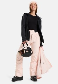 Čierna zip-up bundu, priliehavý čierny top, ružové snehové nohavice s gombíkovým zapínaním, čierne lyžiarske topánky, držiac čiernu prilbu so zrkadlovým zorníkom, svetloružovú bundu.