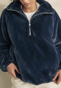 Pulover din fleece bleumarin, cu guler înalt, fermoar pe un sfert și manșete elastice. Textură moale și croială lejeră, potrivit pentru purtarea casual.
