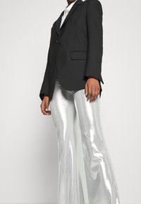 Person wearing a black blazer over a white shirt paired with shiny silver flared pants on a plain light background.