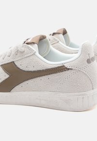 Light grey suede sneakers with a brown leather swoosh and textured white rubber sole. Features distinctive stitching and back tab.