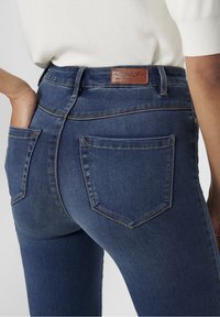 Jeans de mezclilla azul de tiro alto con un corte ajustado, que cuentan con dos bolsillos traseros y un parche de cuero con el logotipo en la cintura. Detalles de costura sutiles.