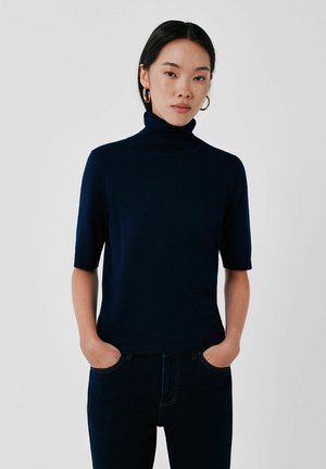 Femme aux cheveux noirs portant un pull à col roulé bleu marine à manches courtes et un jean foncé, debout, les mains dans les poches devant un fond uni.