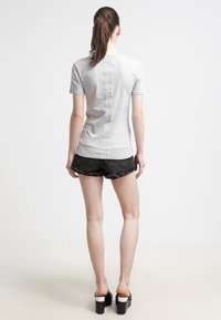 Žena s hnědým ohonem, která nosí světle šedé tričko s krátkým rukávem, černé džíny šortky a černé platformové sandály, stojí zády.