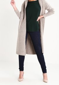 Žena oblečená v béžovém dlouhém kardiganu, tmavě zeleném topu, tmavých úzkých džínech a béžových lodičkách, pózuje s jednou rukou na bok.