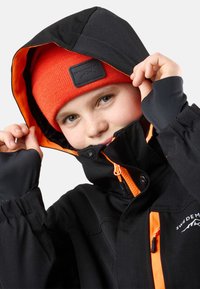 Svart jacka med orange foder och detaljer, med dragkedja fram, huva och elastiska ärmmuddar. Ljusorange stickad mössa på huvudet.