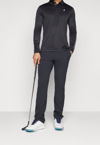 Polo a maniche lunghe nera con una vestibilità slim, abbinata a pantaloni navy e scarpe da golf bianche con dettagli blu, che tiene un bastone da golf.