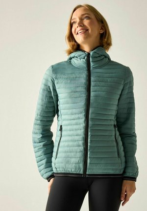 Lachende vrouw die een licht turquoise doorgestikte jas met capuchon en zwarte rits draagt, staande tegen een effen lichte achtergrond, iets omhoog kijkend.