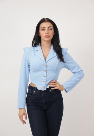 Kobieta z długimi ciemnymi włosami, ubrana w jasnoniebieski krótki blezer z srebrnymi guzikami i ciemne wysokiej talii dżinsy, stoi z jedną ręką na biodrze.
