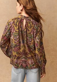 Blusa a fiori color senape con fiori bordeaux, verdi e viola. Presenta una schiena con apertura a chiave e maniche lunghe a sbuffo.
