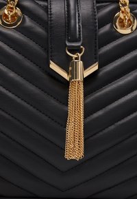 Musta nahkainen käsilaukku, jossa on viivamaista kuviota, kultasävyiset metalliosat ja koristeellinen kultaketju tasseli. Laukussa on jämäkkä muotoilu ja sileä pinta.