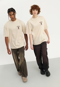 Beige bomulds t-shirts med "Coffee Lovers" grafik, parret med løstsiddende brune denim bukser og træsko. Enkel design, afslappet pasform.