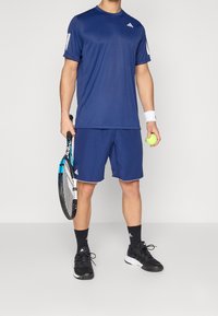 Granatowa koszulka sportowa i szorty z tkaniny odprowadzającej wilgoć. Czarne buty tenisowe, trzymający rakietę tenisową i żółtą piłkę.