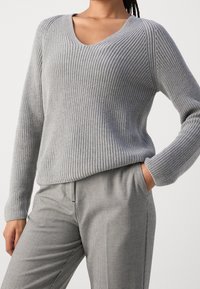 Femme portant un pull en maille côtelée gris clair avec col en V et un pantalon à carreaux gris, main dans la poche, sur fond blanc uni.