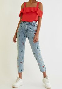 Czerwony top z odkrytymi ramionami i falbanami, zestawiony z jasnoniebieskimi dżinsami z haftem kwiatów i kaktusów oraz białymi trampkami.