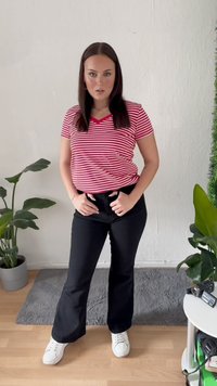 Ženska z dolgimi temnimi lasmi stoji v notranjosti, oblečena v rdečo in belo črtasto majico, črne hlače in bele superge, ob njej pa so rastline in kamera.