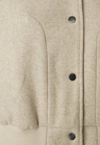 Lekki beżowy płaszcz z mieszanki wełny charakteryzuje się miękką teksturą, czarnymi zatrzaskami oraz dopasowanym ściągaczem u dołu. Wyraźne detale przeszycia na tkaninie.